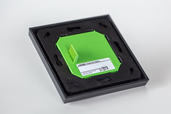 Auf der Rückseite des Loxone Touch for Nano Anthrazit ist das grüne Gehäuse in einer schwarzen Montageplatte verbaut, auf dem die Anschlusskontakte und ein Etikett mit technischen Angaben sichtbar sind.